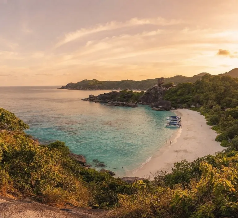 Bay seen from a forested hill at sunset with a boat off a white beach