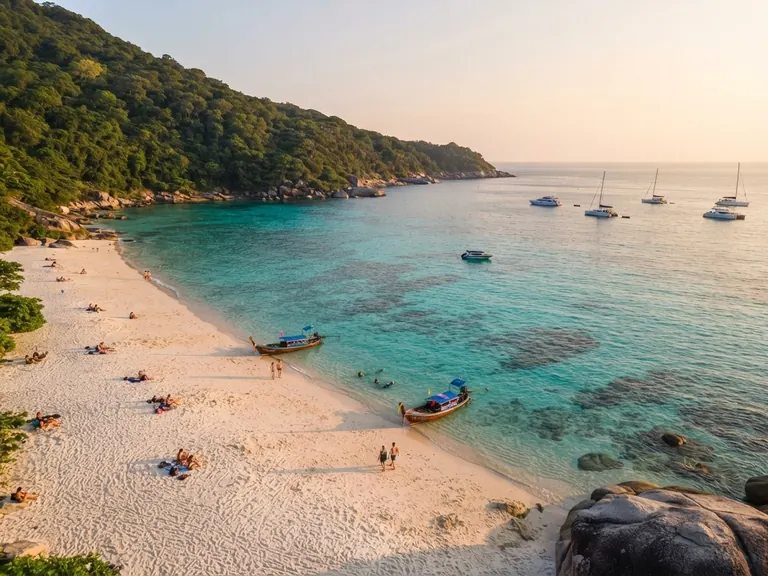 Long white beach with sailboats and longtail boats anchored in a turquoise bay